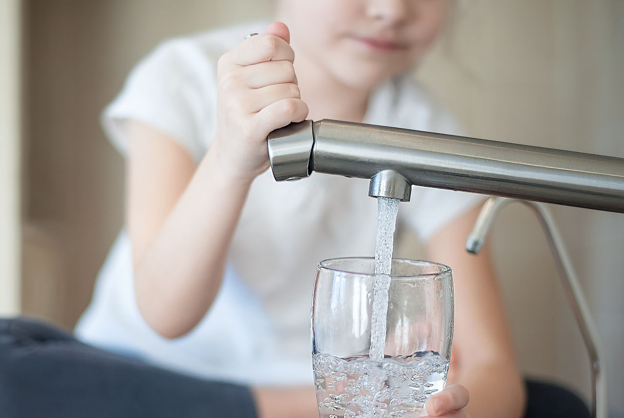 Little girl opens a water tap with her hand holding a transparent glass. Kitchen faucet. Glass of clean water. Filling cup beverage. Pouring fresh drink. Hydration. Healthcare. Healthy lifestyle.