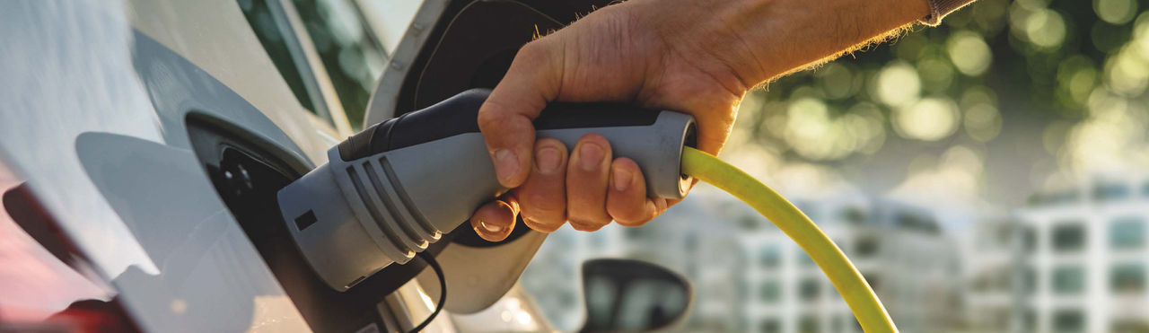 Links im Bild steht ein Elektroauto. Ein Arm reicht von rechts ins Bild und hält den Ladeanschluss zum laden ans Elektroauto.
