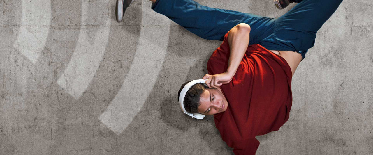 Un homme porte des écouteurs blancs et fait un poirier à un bras. Ses jambes sont pliées en biais et tendues en l'air. En arrière-plan, on voit un mur gris sur lequel sont représentées des lignes blanches en forme d'ondes radio.