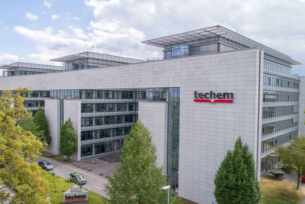 L'image montre un immeuble de bureaux Techem. La façade est claire et se compose de verre et de pierre. Le logo de Techem est bien visible sur le bâtiment. Devant le bâtiment se trouvent plusieurs arbres et deux voitures. Le ciel est en partie nuageux.