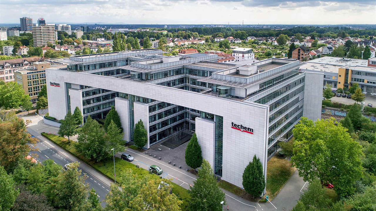 L'image montre un immeuble de bureaux Techem. La façade est claire et se compose de verre et de pierre. Le logo de Techem est bien visible sur le bâtiment. Devant le bâtiment se trouvent plusieurs arbres et deux voitures. Le ciel est en partie nuageux.