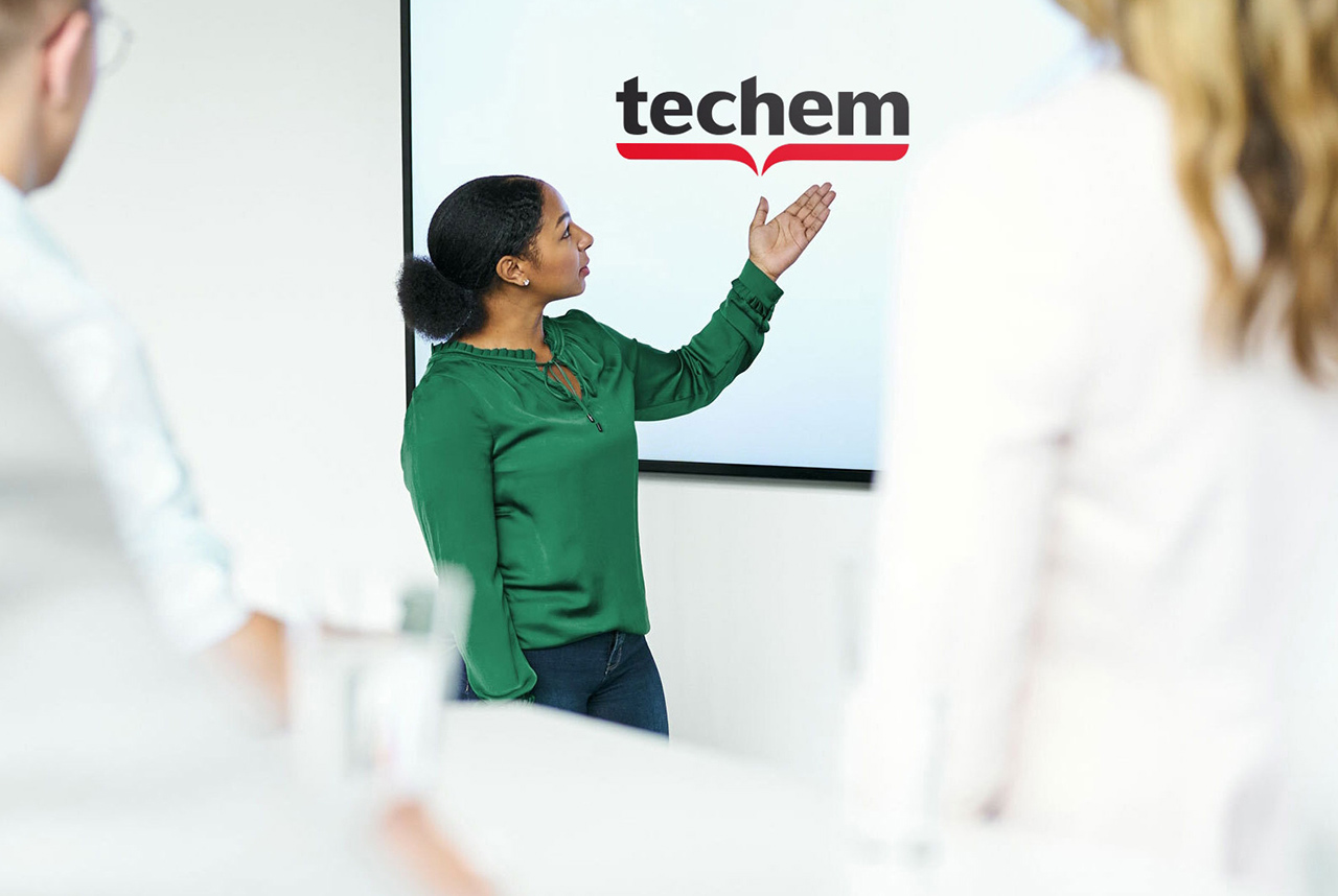 Dans une salle de réunion, une femme en chemisier vert fait une présentation devant un écran portant le logo de Techem. Deux personnes se tiennent au premier plan et écoutent attentivement.
