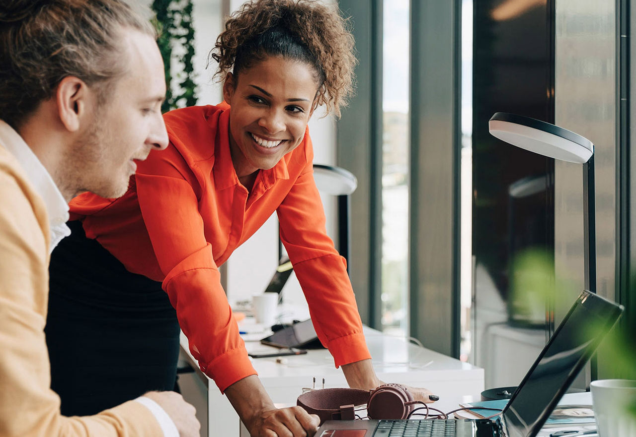 Des collègues de travail souriants : une femme debout en chemise orange et un homme assis en pull brun clair regardent ensemble un ordinateur portable sur leur lieu de travail.