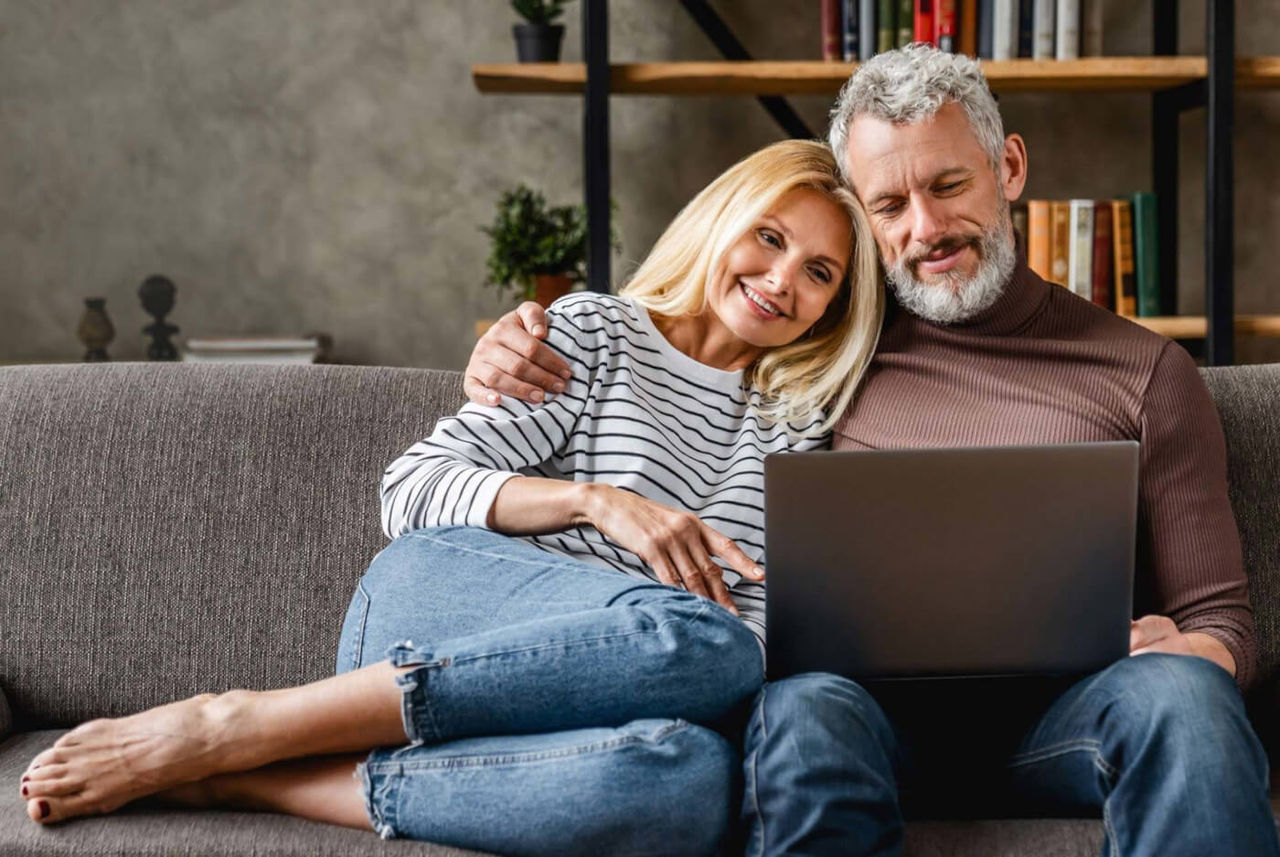 Ein glückliches Paar sitzt gemütlich auf dem Sofa und schaut gemeinsam auf einen Laptop.