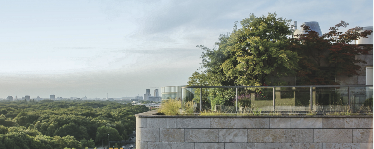 Moderne Dachterrasse mit Glasgeländer, Pflanzen und Bäumen, mit Blick auf die Stadt und einen grünen Park.