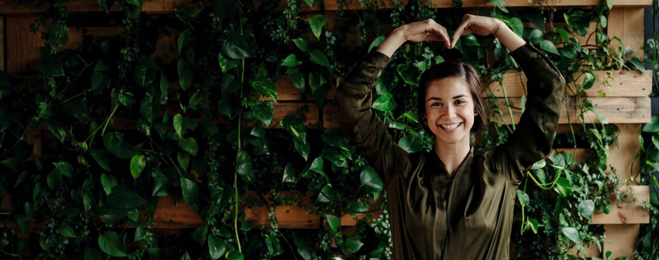  Frau hebt die Arme über den Kopf und formt ein Herz mit ihren Händen, während sie lächelt.