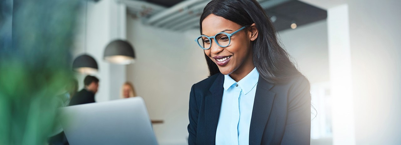 Eine junge Frau mit einer hellblauen Brille sitzt im Büro vor einem Laptop und lächelt. Im Hintergrund sieht man weitere zwei Kollegen.